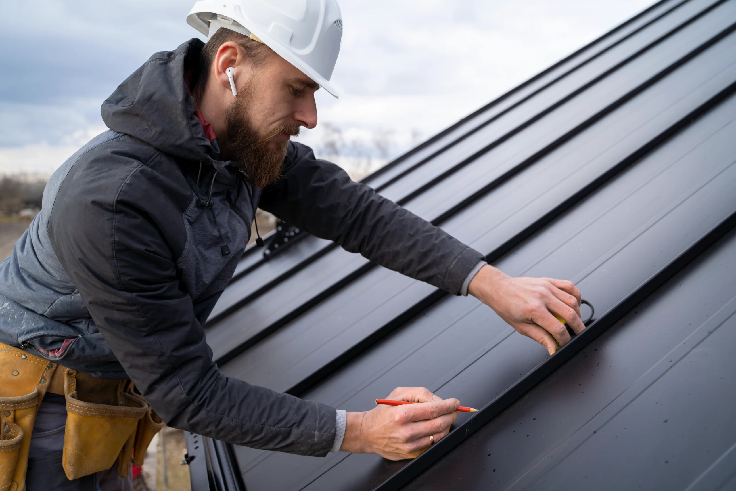 Roofer in a hard hat measuring black standing seam metal roofing with a tape measure and red pencil.