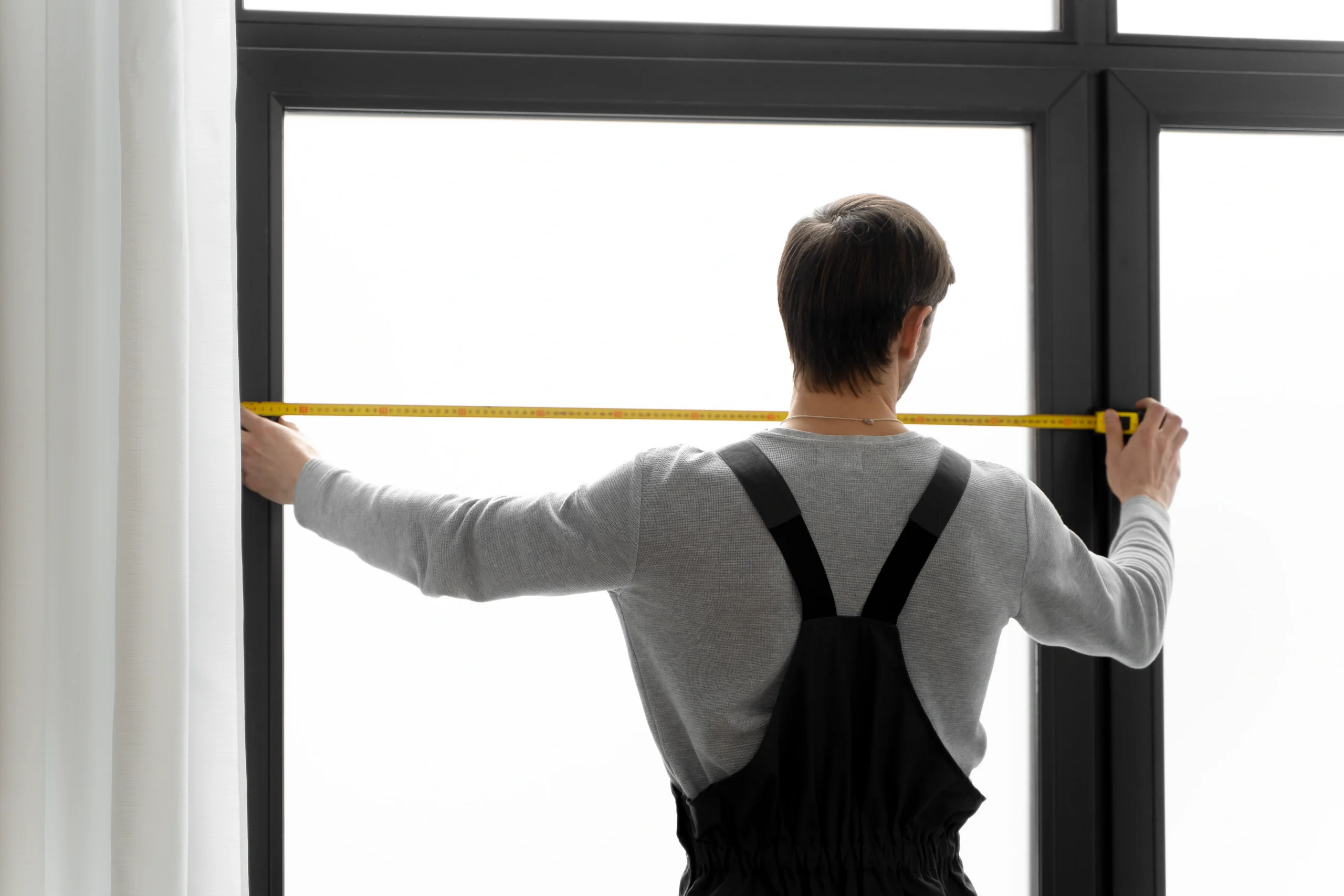 Worker in black overalls measuring a large black-framed window with a yellow tape measure, seen from behind.